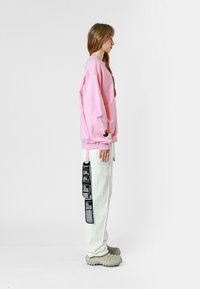 Sudadera rosa, pantalones blancos con etiquetas negras y zapatos gris texturizados. Corte relajado y cordón visible en los pantalones.