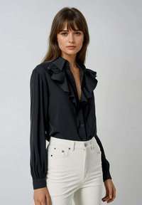 Blouse noire avec des détails froncés le long du col et de la patte de boutonnage, manches longues avec des poignets froncés, associée à un jean blanc taille haute.