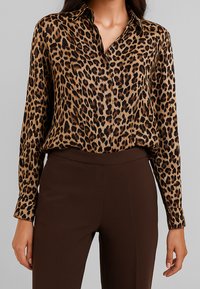 Blusa in stampa leopardata con colletto e chiusura a bottoni, dotata di maniche lunghe e una texture di tessuto morbida e liscia. Abbinata a pantaloni marroni.