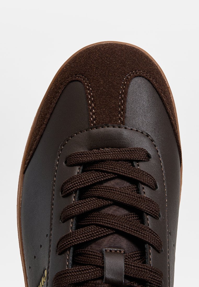 Braune Leder-Sneaker mit einer Wildleder-Zehenkappe, braunen Schnürsenkeln und einer strukturierten Gummisohle. Nähedetails sind an den Kanten sichtbar.