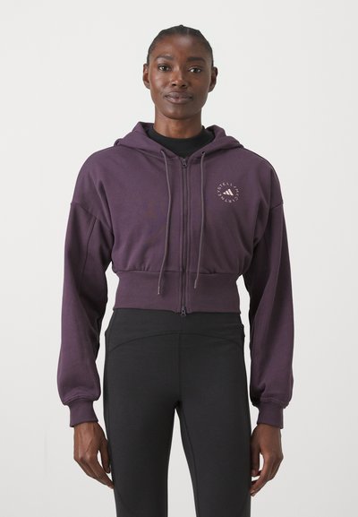 Woman wearing a cropped purple zip-up hoodie with logo on chest and black high-waisted leggings against a plain background.