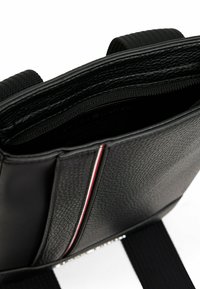 Interior de un bolso de cuero texturizado negro con un bolsillo con cremallera, que presenta una franja en rojo y blanco a lo largo del borde y correas negras ajustables.