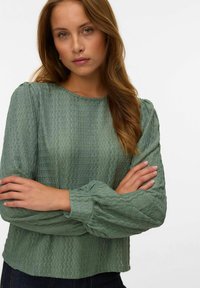 Femme jeune avec des cheveux châtain clair portant un blouse à manches longues texturée de couleur vert sauge, bras croisés, expression neutre, fond blanc.