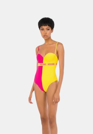 Donna in piedi che indossa un costume intero color-block rosa e giallo con spalline sottili e fascia con logo Moschino in vita.