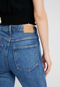 Jean denim bleu taille haute avec un patch en cuir à l'arrière, comprenant deux poches arrière et des coutures visibles. Texture lisse.
