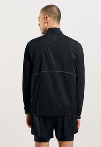 Schwarze, leichte Jacke mit durchgehendem Reißverschluss, hohem Kragen und reflektierenden Akzenten. Die Rückseite verfügt über ein Logo und durchbrochene Details.