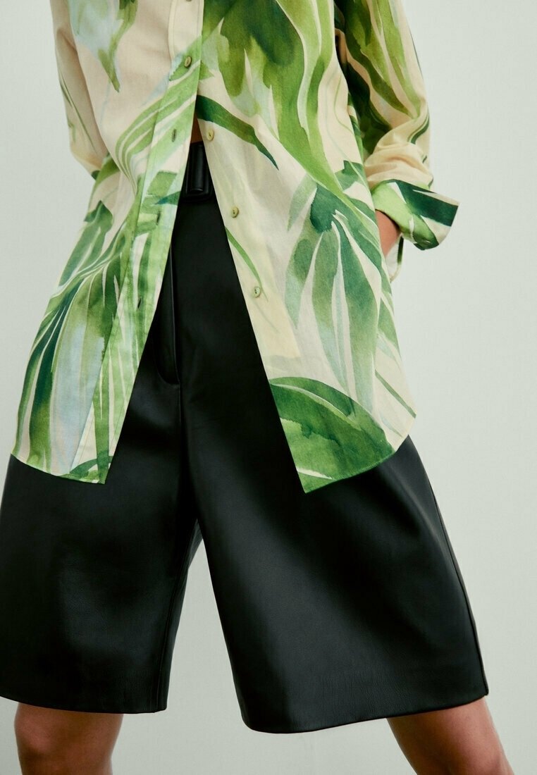 Personne portant une chemise à motifs feuilles vert et crème rentrée dans un short en cuir noir ample mi-genoux, mains dans les poches.