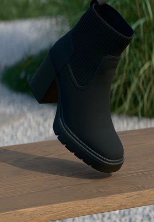 Bottine noire à talon épais avec tige en maille côtelée, posée sur une surface en bois avec un fond de verdure.