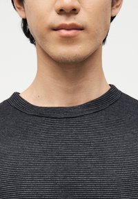 Νεαρό άτομο με ελαφριά φυτρωμένα γένια, φορούσε σκούρα γκρι μπλούζα με λεπτές οριζόντιες ρίγες, μπροστά σε ένα απλό φόντο.