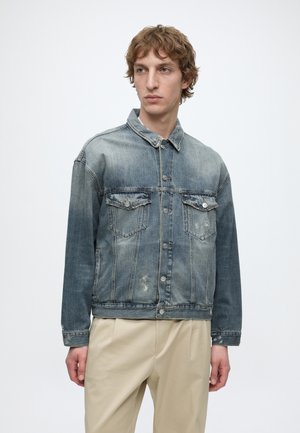 Mladý muž nosiaci vyblednutú modrú rifľovú bundu s zapínateľnými vreckami na hrudi a béžové nohavice, stojaci pred jednoduchým bielym pozadím.