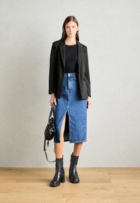 Schwarze, taillierte Blazer über einem schwarzen Top, kombiniert mit einem blauen Jeans-Bleistiftrock mit einem vorderen Schlitz. Schwarze Stiefel mit Absatz und eine schwarze Handtasche.