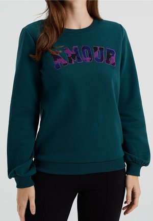 Femme portant un sweat-shirt à manches longues vert foncé avec un texte "AMOUR" à motifs violets et roses sur la poitrine, associé à un pantalon noir.