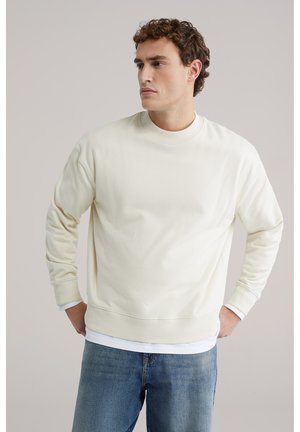 Lichtbeige sweatshirt, ronde hals, lange mouwen, geribbelde boorden en zoom, zachte textuur. Gedragen over een witte longsleeve met blauwe spijkerbroek.