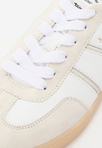 Sneaker bianca e crema realizzata in materiali scamosciati e in pelle, con punta rotonda, lacci bianchi, cuciture visibili e suola in gomma color caramello.