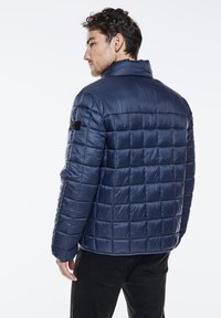 Navy blue puffer jacket with a grid pattern, high collar, and a black label detail on the left sleeve. Soft, shiny material.