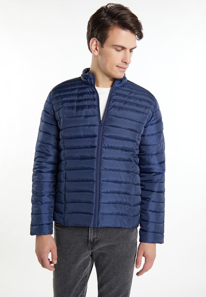 Giacca imbottita blu navy con cuciture orizzontali, colletto alto e chiusura con zip. Realizzata in materiale lucido, abbinata a jeans grigi.