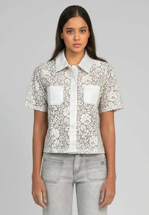 Jeune femme portant une chemise à manches courtes en dentelle florale blanche avec col uni et poches avant, associée à un jean gris clair.