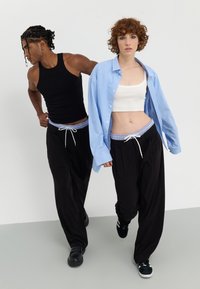 Pantalons noirs amples avec taille élastique, portés avec des crop tops blancs et une chemise boutonnée rayée bleu clair. Des baskets complètent le look.