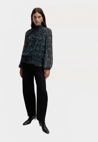 Fließende blaue Bluse mit Blumenmuster, hohem Kragen und langen transparenten Ärmeln, kombiniert mit schwarzen Hosen und Knöchelboots.