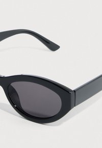 Μαύρα γυαλιά ηλίου με οβάλ φακούς, λεία πλαστική σκελετό και φαρδιά πλευρικά μπράτσα. Οι φακοί είναι σκουρόχρωμοι γκρι, προσφέροντας πλήρη κάλυψη.