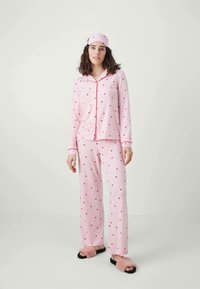 Rosa pyjamasset med röd hjärmönster, som har långa ärmar, skjorta med knappar och matchande byxor; mjukt tyg; inkluderar en matchande sovmask.