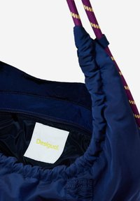 Dunkelblaues Tasche mit einer weichen, glänzenden Textur, versehen mit einem gerafften oberen Rand und lila Griffen mit gelben Akzenten. Das Etikett trägt die Aufschrift "Desigual."