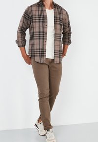 Hnědá kostkovaná flanelová košile přes krémové tričko s kulatým výstřihem, spárovaná s hnědými slim-fit kalhotami a bílými teniskami, předvádějící ležérní vzhled.