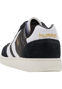 Sapatilha de camurça e couro preta com detalhes brancos e logotipo dourado "hummel". Apresenta três riscas brancas e uma sola branca texturizada.