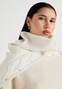 Κρέμα oversized πο�υλόβερ με ριμπ υψηλό λαιμό, που διαθέτει λεπτομέρειες από καλώδιο και κουμπιά στον ώμο. Μαλακή υφή.