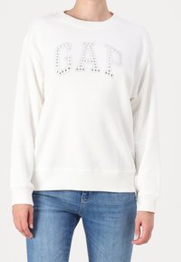 Persona che indossa un felpa bianca con lettere "GAP" in metallo argentato e jeans blu, in piedi contro uno sfondo chiaro e uniforme.