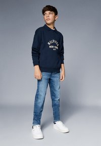Paauglys stovi, vilkintis tamsiai mėlyną ilgomis rankovėmis marškinį su užrašu "Hilfiger New York City", mėlynas džinsus ir baltas sportines batus, žiūrintis šoną.