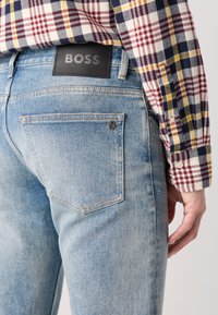 Light blue denim jeans with a black leather "BOSS" label on the back waistband. Features subtle stitching and a back pocket with rivet detail.