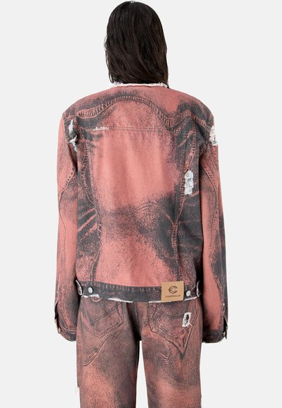 Personne aux longs cheveux bruns portant une veste en jean délavée rose pâle et noire usée, ainsi qu'un pantalon assorti, vue de dos.