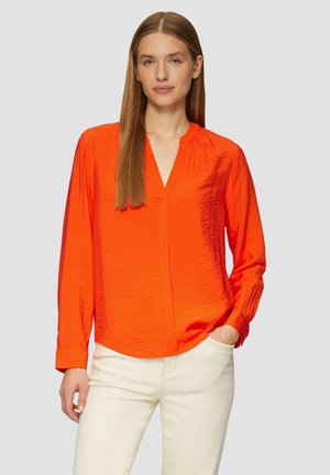 Falte Bluse in Orange mit V-Ausschnitt, langen Ärmeln und Raffung an der Schulter; kombiniert mit hellem Jeans.