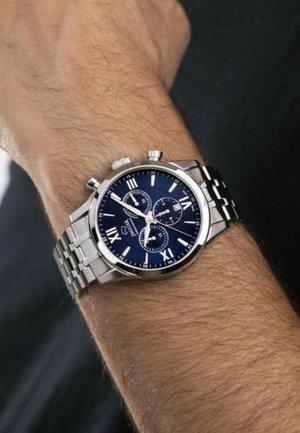 Montre-bracelet chronographe avec un bracelet en acier inoxydable, un cadran bleu, des accents argentés, des chiffres romains et des sous-cadrans pour des fonctionnalités supplémentaires.