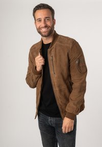 Giacca bomber in suede marrone con polsini e colletto a coste, dotata di una zip frontale e di una tasca con zip sulla manica sinistra. T-shirt nera sotto.