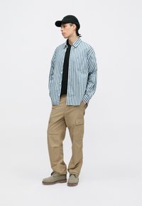 Prúžkovaná modro-biela košeľa s gombíkmi, čierny tričko s okrúhlym výstrihom, khaki cargo nohavice a šedé topánky s čiernou čiapkou. Neformálny outfit s vreckami.