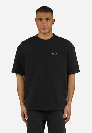 Fiatal férfi rövid hajjal, fekete, túlméretezett pólót visel, amelyen fehér "Pegador" felirat található a mellkasán, egyszínű háttér előtt áll.