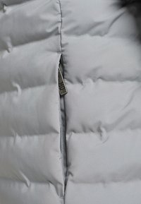 Sezione di un giubbotto a vento grigio con una texture liscia e leggermente lucida. Tasca con zip molto evidente dotata di un cursore metallico.
