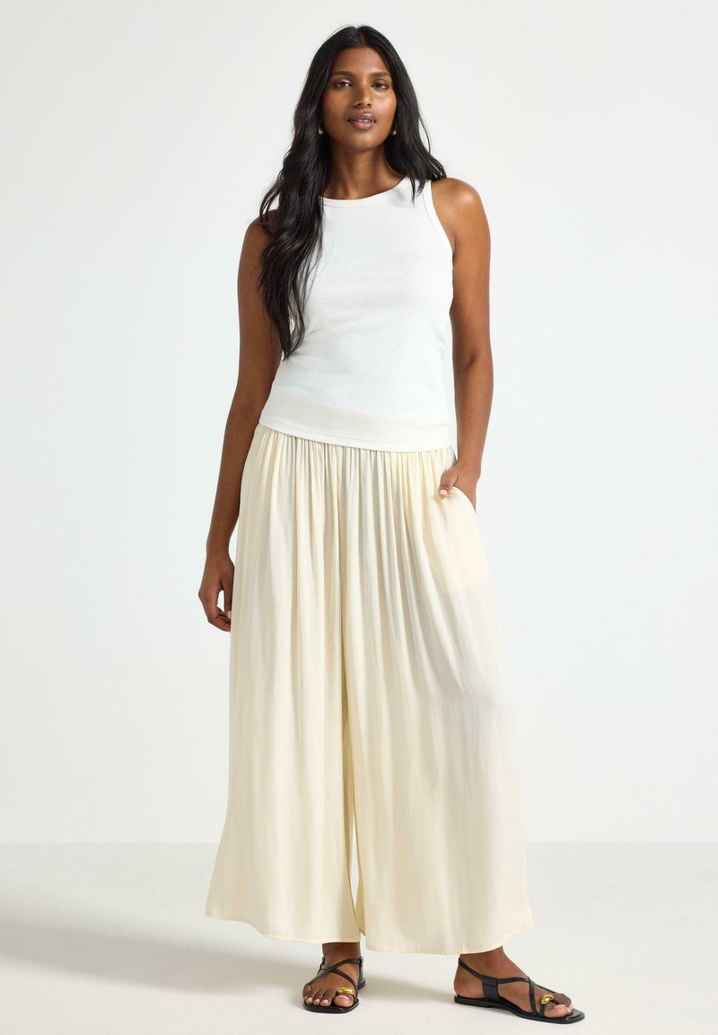 Top bianco senza maniche abbinato a pantaloni palazzo larghi di colore crema. I pantaloni presentano una vita arricciata e un tessuto leggero e morbido. Sandali neri.