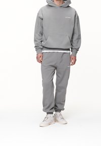 Felpa grigia con cappuccio e tasca frontale, abbinata a pantaloni jogger coordinati. Entrambi presentano una texture morbida e un sottile dettaglio del logo.