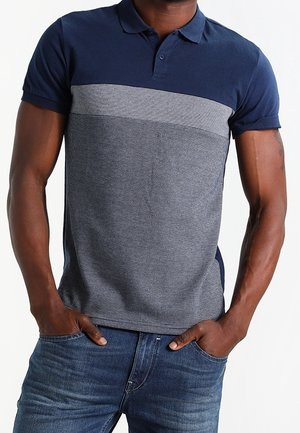 Man draagt een donkerblauw en grijs polohemd met korte mouwen, gecombineerd met een blauwe spijkerbroek, staand met handen in de zakken tegen een witte achtergrond.