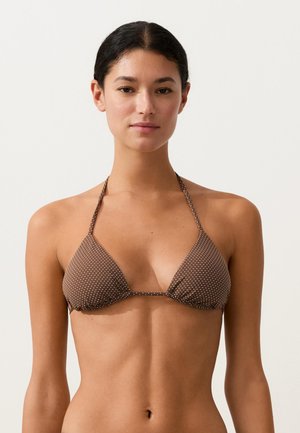 Junge Frau mit dunklen, zurückgebundenen Haaren, die ein braunes Dreieck-Bikinioberteil mit Punkten trägt, blickt nach vorne vor einem schlichten, hellen Hintergrund.