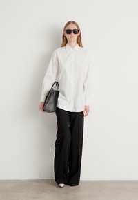 Camicia bianca con bottoni, pantaloni neri con gamba ampia, borsa nera e occhiali da sole. Tessuto morbido e design su misura. Colori neutri, estetica moderna.