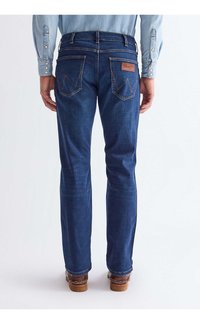 Muž nosi tmavomodré rovné rifle s zadnými vreckami a hnedými koženými topánkami, stojaci na jednoduchom svetlom pozadí.