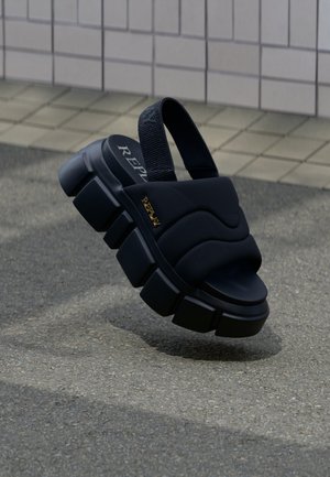 Chunky black sandal with thick sole, padded straps, and a heel sling on grey pavement against tiled wall.
