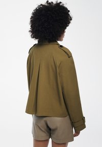 Giacca verde oliva con retro plissettato e spalline sulle spalle. Il materiale sembra liscio, i polsini hanno dettagli con bottoni. Indossata sopra pantaloni corti chiari.