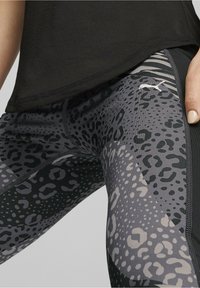 Nahaufnahme einer Person in leggings mit schwarzem und grauem Leopardenmuster und Puma-Logo, kombiniert mit einem schwarzen Oberteil und der Hand auf der Hüfte.