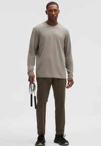 Długi rękaw, jasnoszary top o luźnym kroju, zestawiony z oliwkowozielonymi spodniami. Męski model trzyma czarną skakankę i nosi czarne trampki.