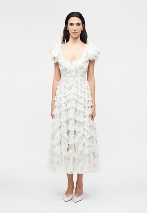 Γυναίκα με μακριά σκούρα μαλλιά που φοράει λευκό στεφανωτό midi φόρεμα με μανίκια φουσκωτά και άσπρα μυτερά τακούνια, στέκεται σε λευκό φόντο.